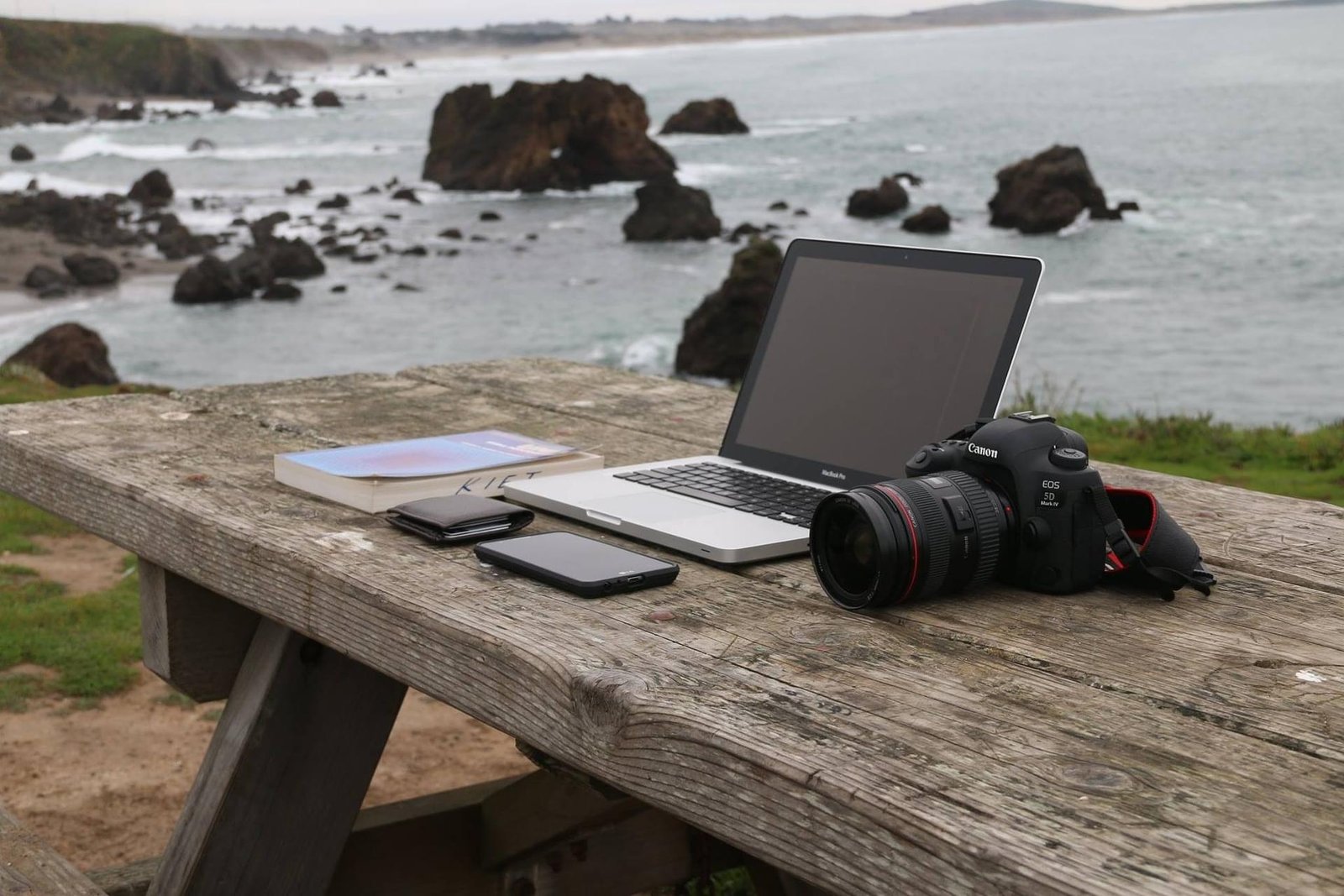 Laptop, phone, book and camera lying on a wooden table. In the background looms a rocky coastline.